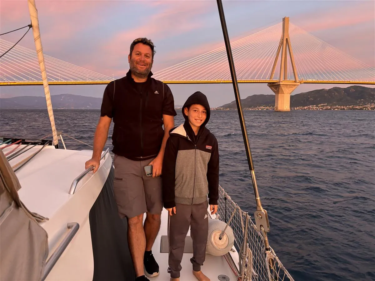 Shai on the bow of a catamaran at sunset, the Rio-Antirrio bridge behind him