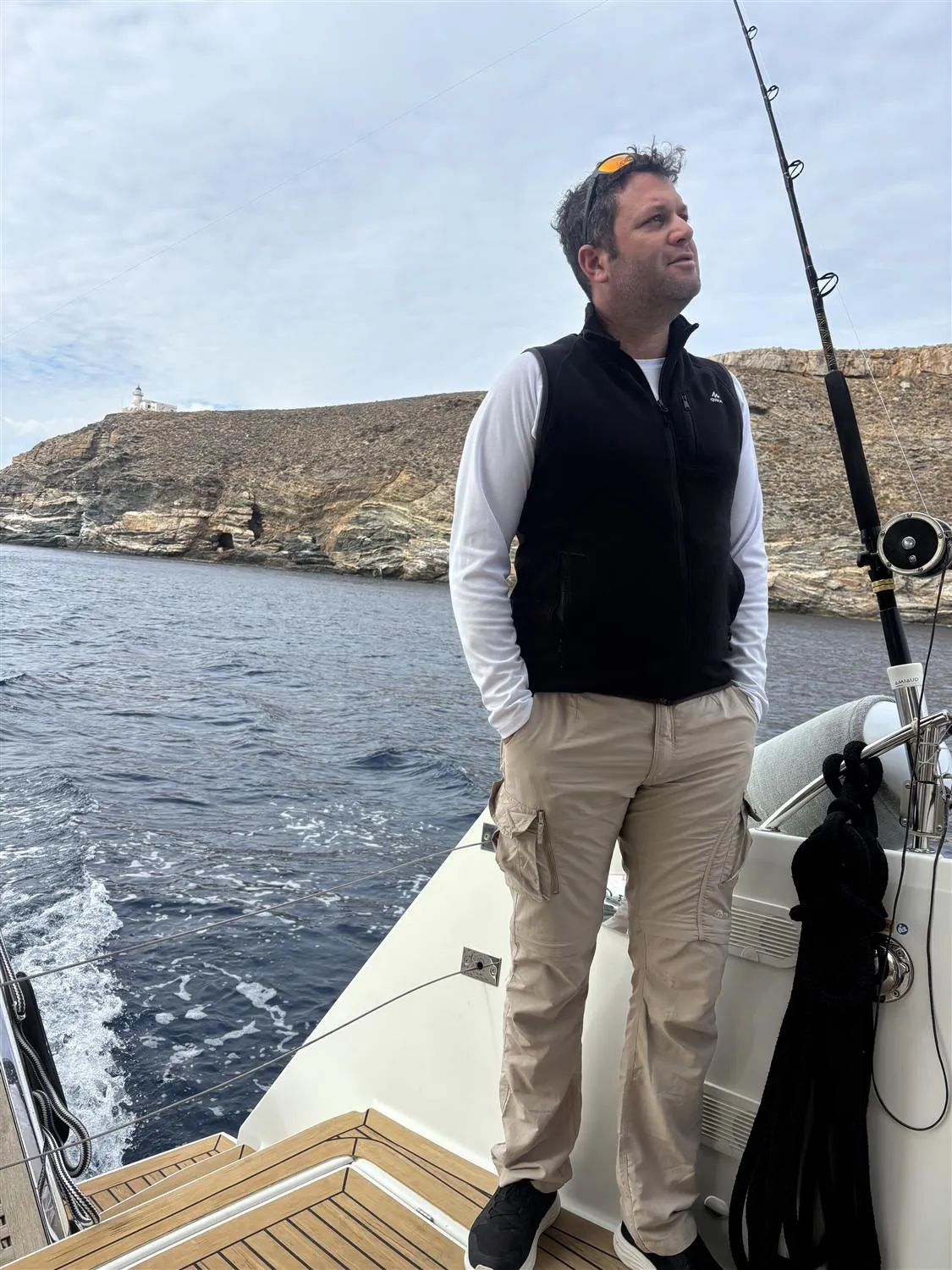Shai Tilinger on the aft deck of a sailing yacht, a headland and lighthouse behind him
